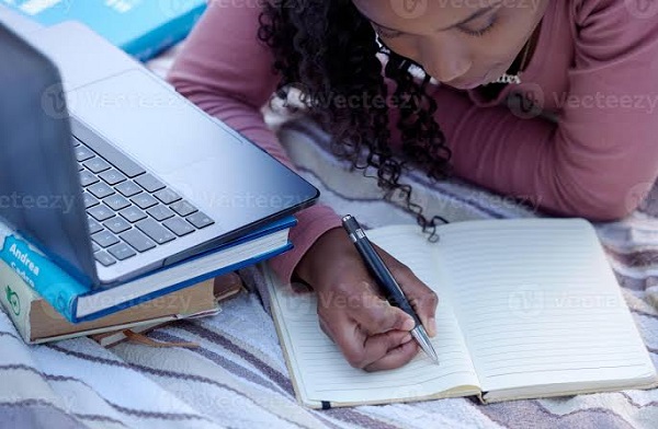 Student in a library taking notes while researching on a laptop